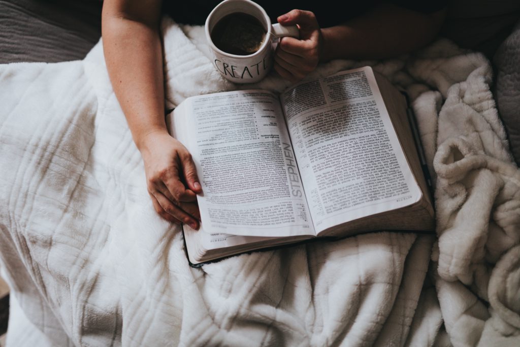 A person sits in bed with a cup of coffee and an open Bible, praying a morning prayer for calm.